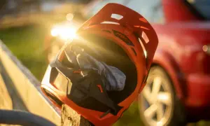 En orange mx hjälm med en grå trasa inuti vilar på ett trästaket. Solen skiner genom hjälmen, och en röd bil syns i bakgrunden.