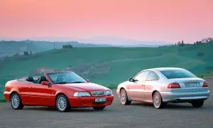 Två Volvo C70, en röd cabriolet och en silverfärgad coupé, står på en grusväg med böljande gröna kullar och en rosa himmel i bakgrunden. En volvo c70 cab problem kan vara att taket inte fälls ner.