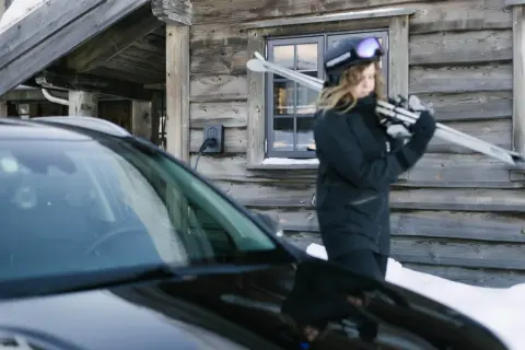 En person med skidor laddar sin elbil vintertid vid en timmerstuga.