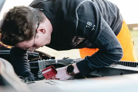 Man med glasögon böjd över bilmotorn, kanske för att fixa det brummande ljudet från bilen.