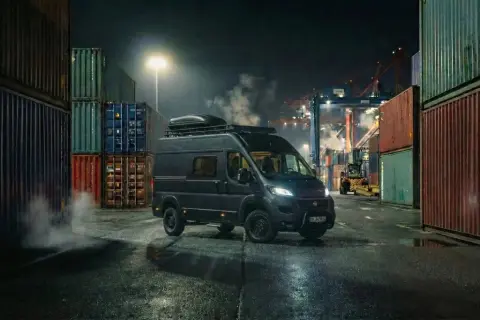 Mörk Fiat Ducato står vid en hamn, omgiven av containrar. Rök stiger från fordonet, vilket antyder ett fiat ducato motor problem.