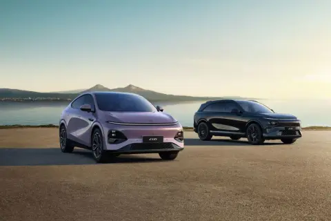 Två XPeng SUV:ar, en lila och en svart, står vid en sjö med berg i bakgrunden.