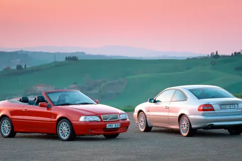 Två Volvo C70, en röd cabriolet och en silverfärgad coupé, står på en grusväg med böljande gröna kullar och en rosa himmel i bakgrunden. En volvo c70 cab problem kan vara att taket inte fälls ner.