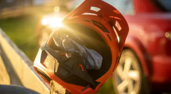 En orange mx hjälm med en grå trasa inuti vilar på ett trästaket. Solen skiner genom hjälmen, och en röd bil syns i bakgrunden.