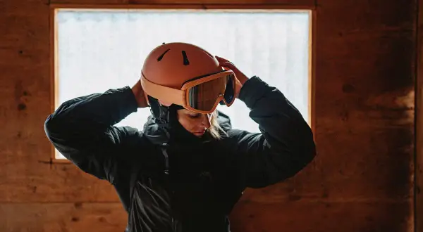 Kvinna justerar sin orangea suomy hjälm och skidglasögon framför ett fönster i en trästuga.