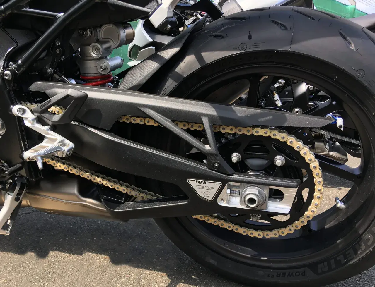 Detaljbild på en motorcykels bakparti med ett gyllene kedjespel mc som löper över ett svart drev. Däcket är ett Michelin.