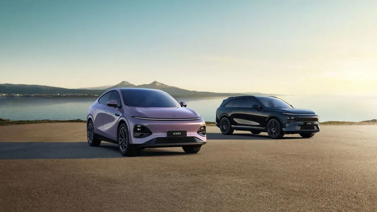Två XPeng SUV:ar, en lila och en svart, står vid en sjö med berg i bakgrunden.