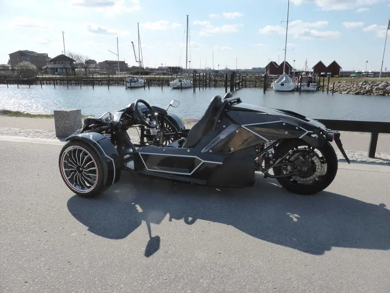 Svart, futuristisk motorcykel på b-kort står vid en hamn med segelbåtar.