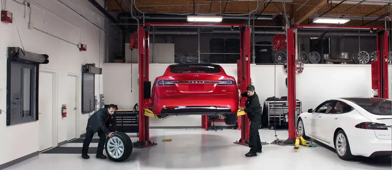 Mekaniker utför service på en röd Tesla Model 3 som lyfts i en verkstad. En vit Tesla Model 3 står bredvid.