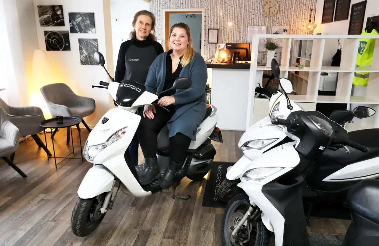 Tv&aring; kvinnor sitter p&aring; en vit moped. Kanske de diskuterar mopedkort pris och hur man f&aring;r det.