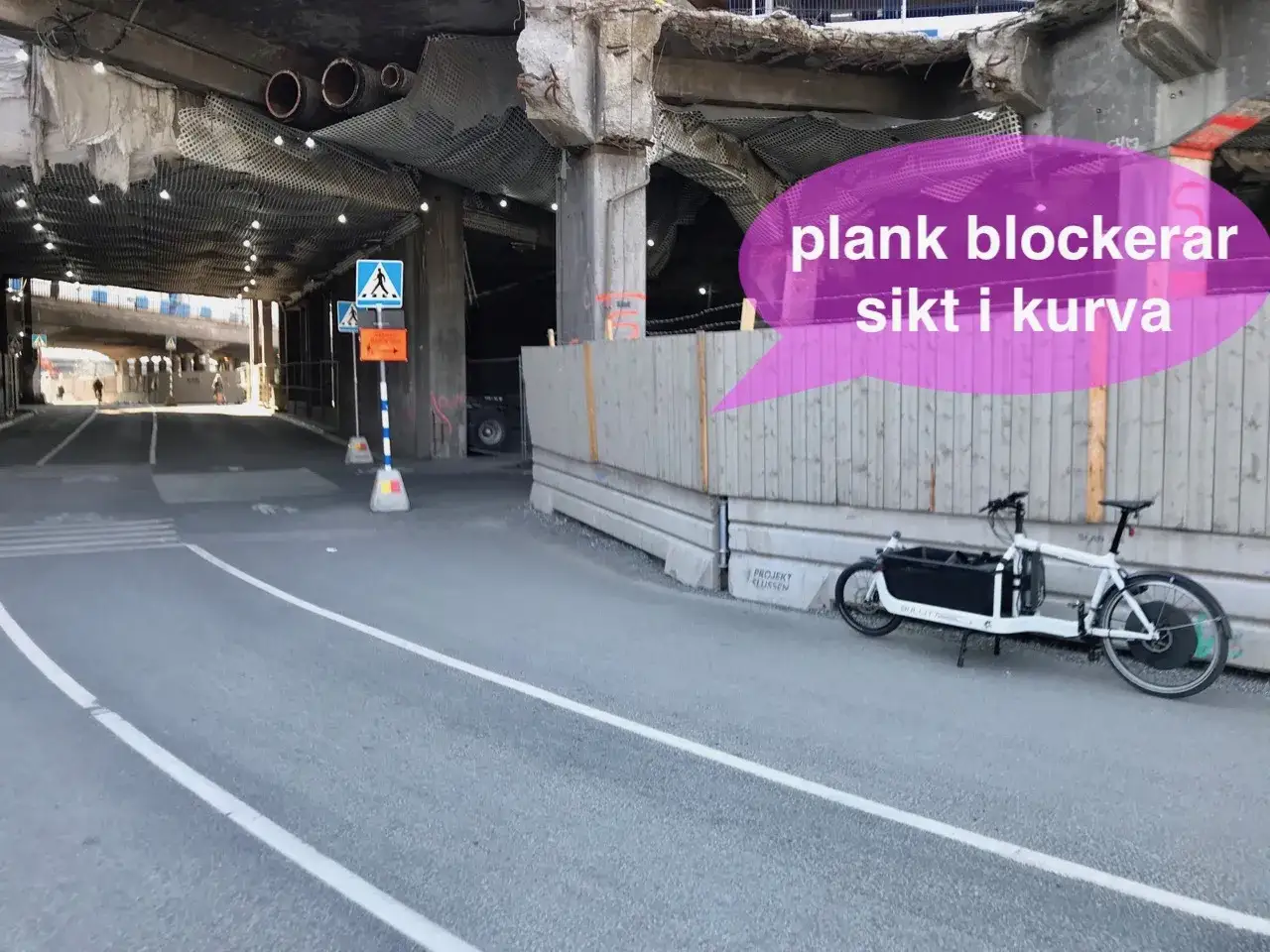 En lastcykel st&aring;r parkerad vid en v&auml;g under en bro. Plankor blockerar sikt i kurvan, vilket skapar en k&auml;nsla av dold fara.