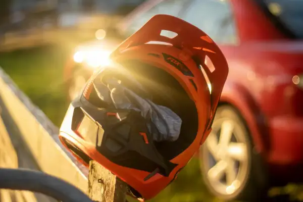 En orange mx hjälm med en grå trasa inuti vilar på ett trästaket. Solen skiner genom hjälmen, och en röd bil syns i bakgrunden.