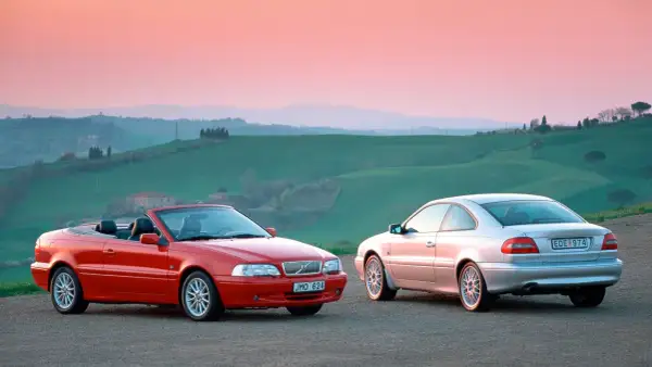 Två Volvo C70, en röd cabriolet och en silverfärgad coupé, står på en grusväg med böljande gröna kullar och en rosa himmel i bakgrunden. En volvo c70 cab problem kan vara att taket inte fälls ner.